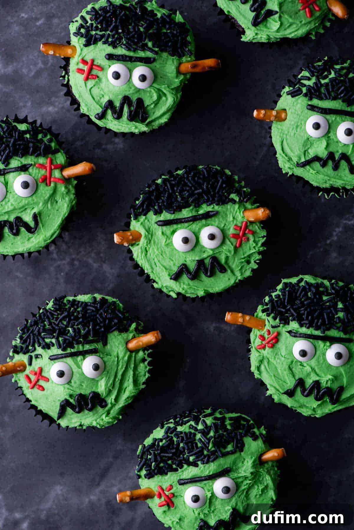 Frankenstein cupcakes displayed on a grey surface, showcasing their green frosting, candy eyes, black hair sprinkles, red stitches, and pretzel stick bolts.