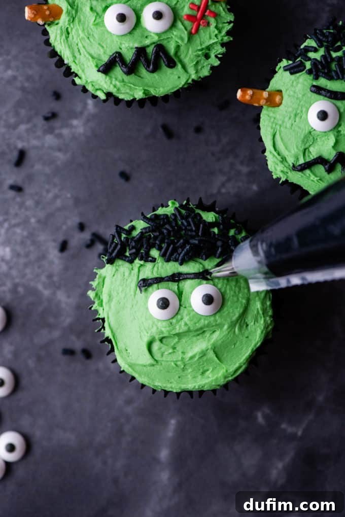 A Frankenstein cupcake in the process of being decorated with piped black eyebrows, shown alongside other completed cupcakes.