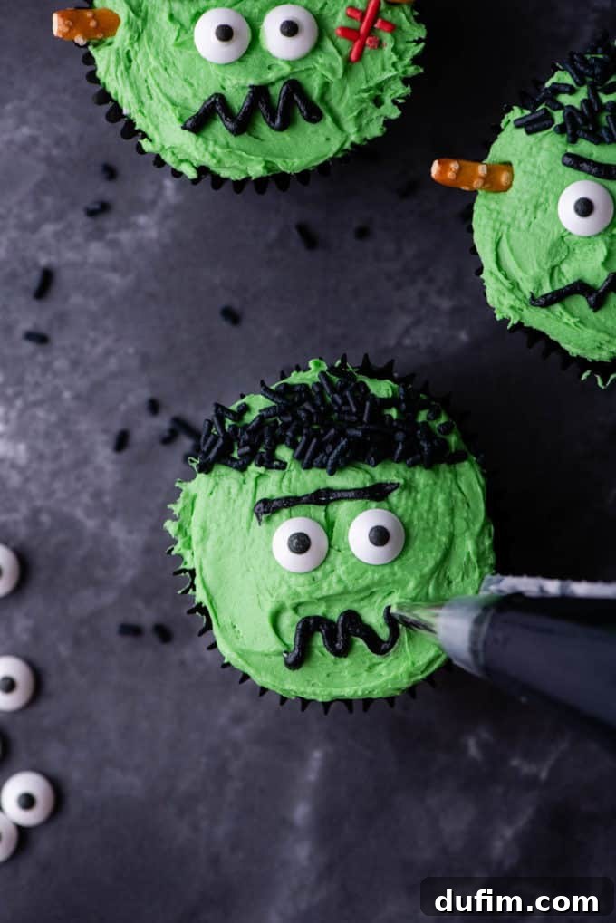 A Frankenstein cupcake being decorated with a piped black squiggly mouth, next to finished Frankenstein cupcakes.