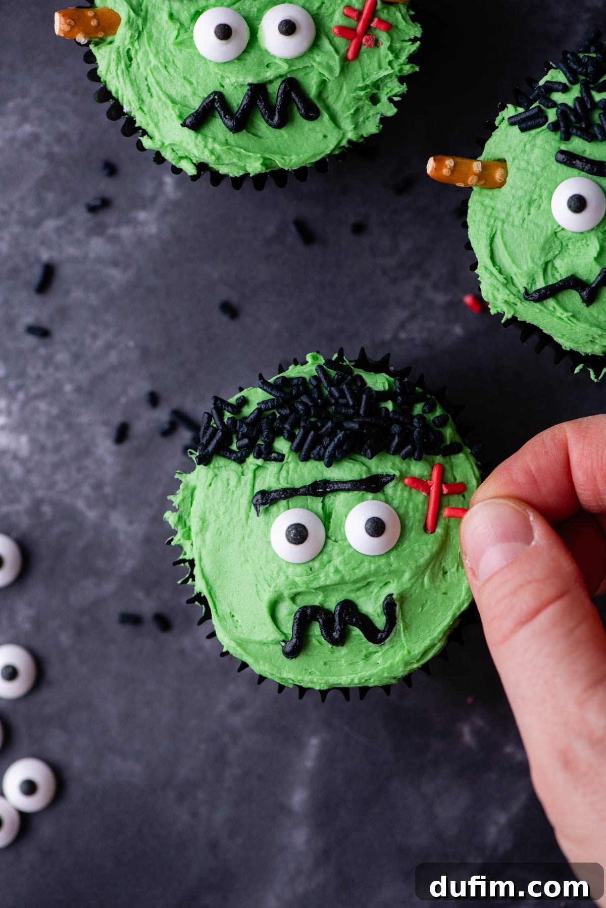 A Frankenstein cupcake being adorned with red sprinkles to form the stitches, surrounded by other complete Frankenstein cupcakes.