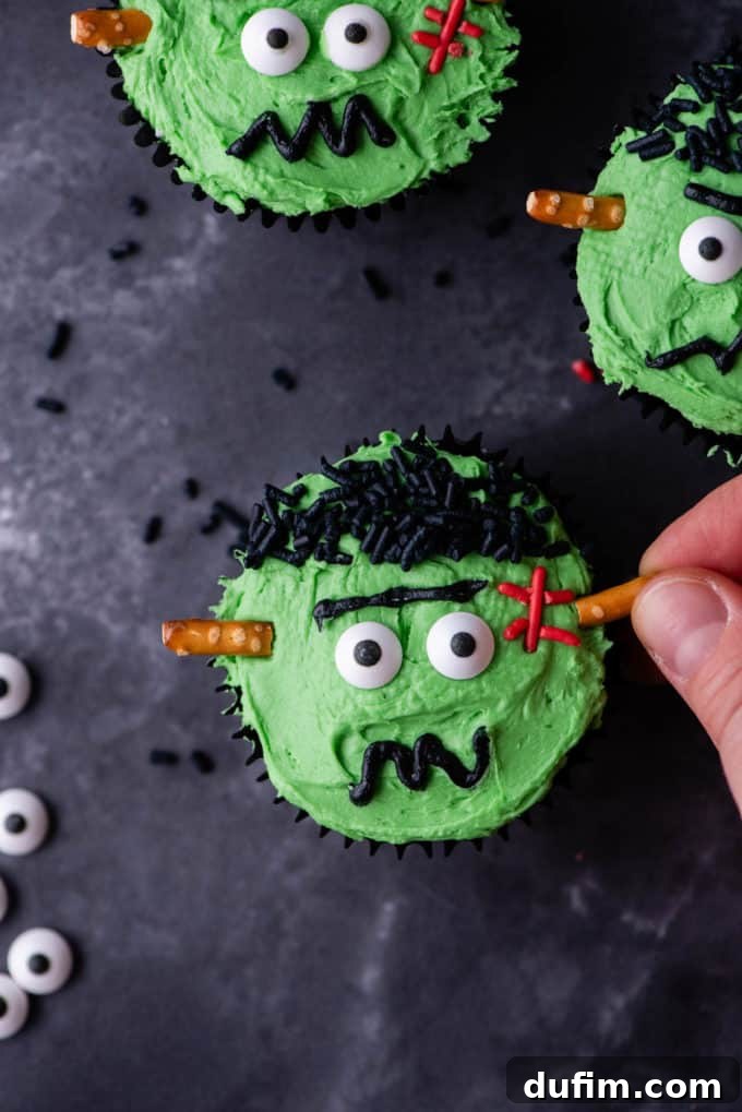 A Frankenstein cupcake being completed by inserting pretzel sticks as neck bolts, alongside finished cupcakes.