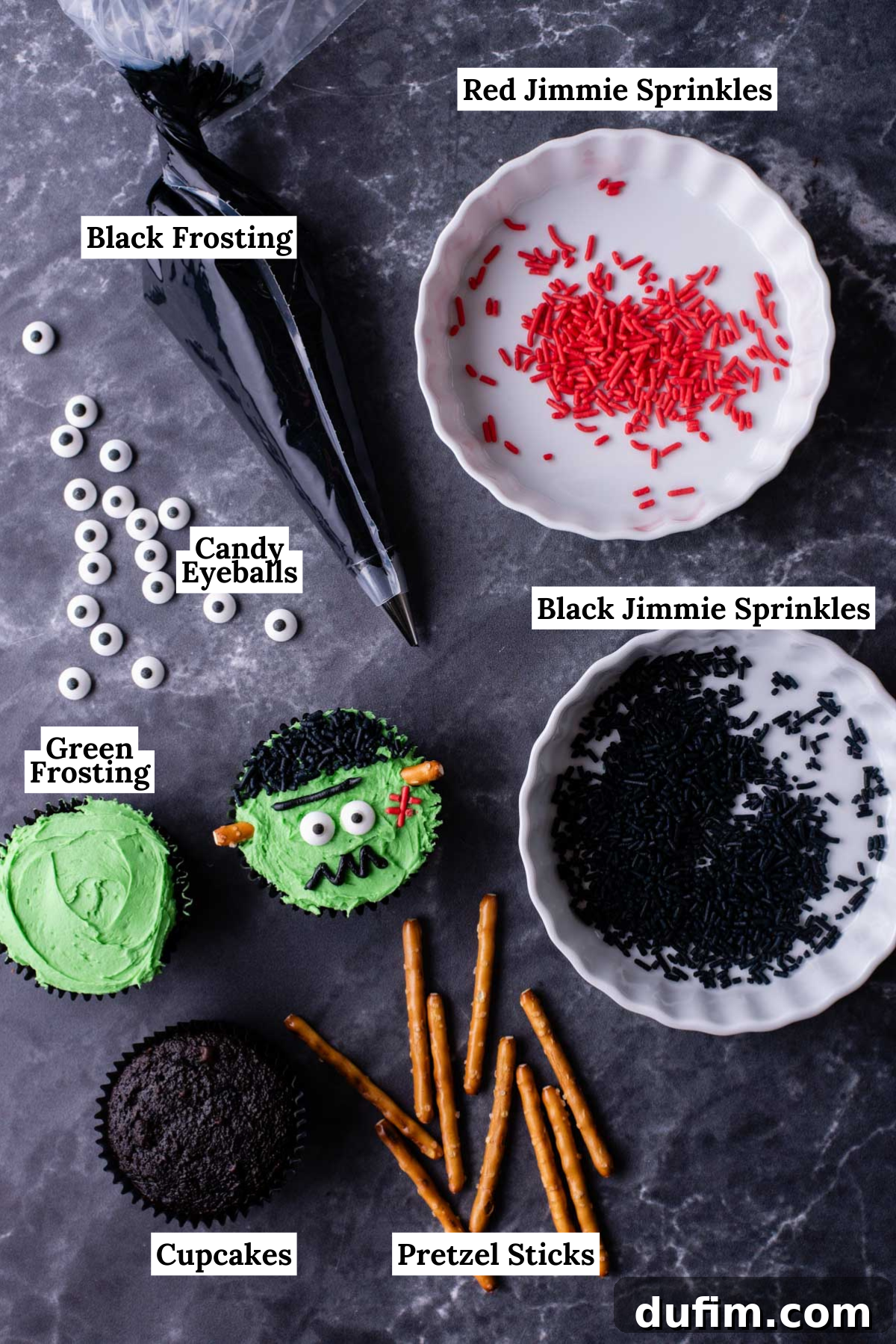 Ingredients laid out for Frankenstein cupcakes, including red jimmie sprinkles, black jimmie sprinkles, baked chocolate cupcakes, pretzel sticks, candy eyeballs, and black frosting, ready for assembly.