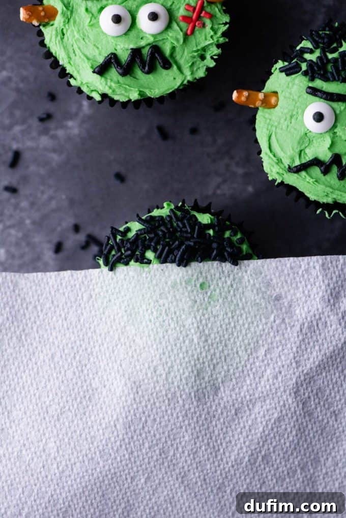 A cupcake being prepared with green frosting, partially covered by paper towel for applying black sprinkles, next to other completed Frankenstein cupcakes.