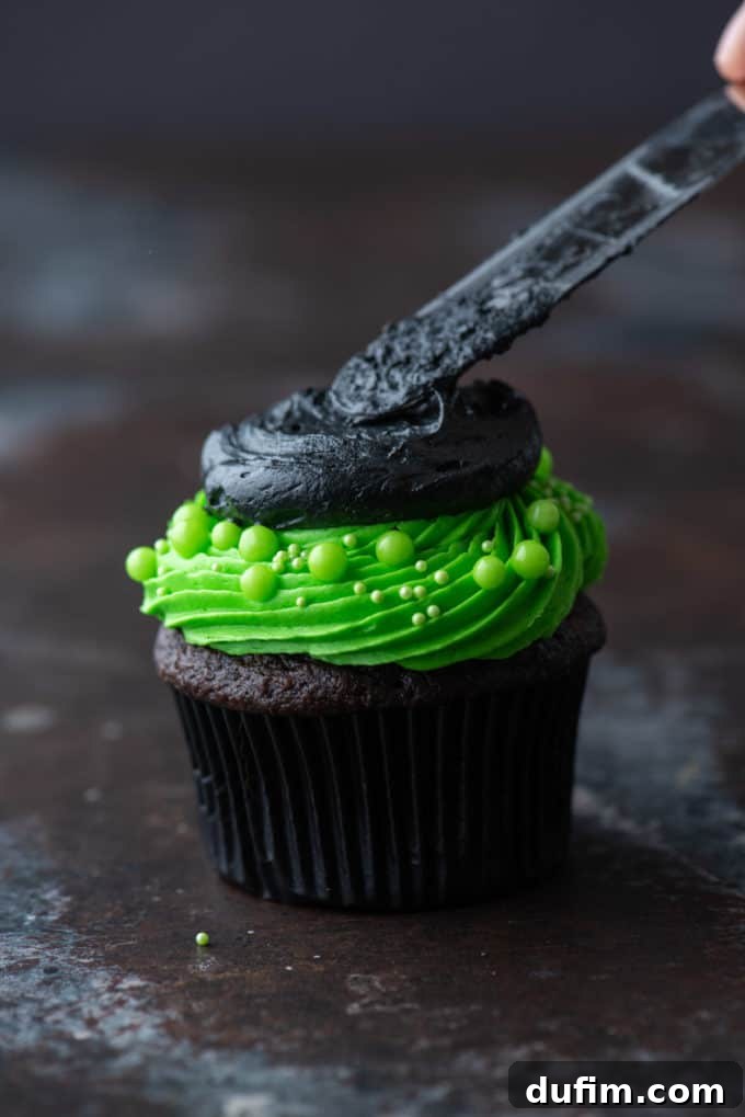 A chocolate cupcake with bright green frosting and a black frosting circle on top, being gently spread and shaped with a small knife to create a smooth, defined witch hat brim.