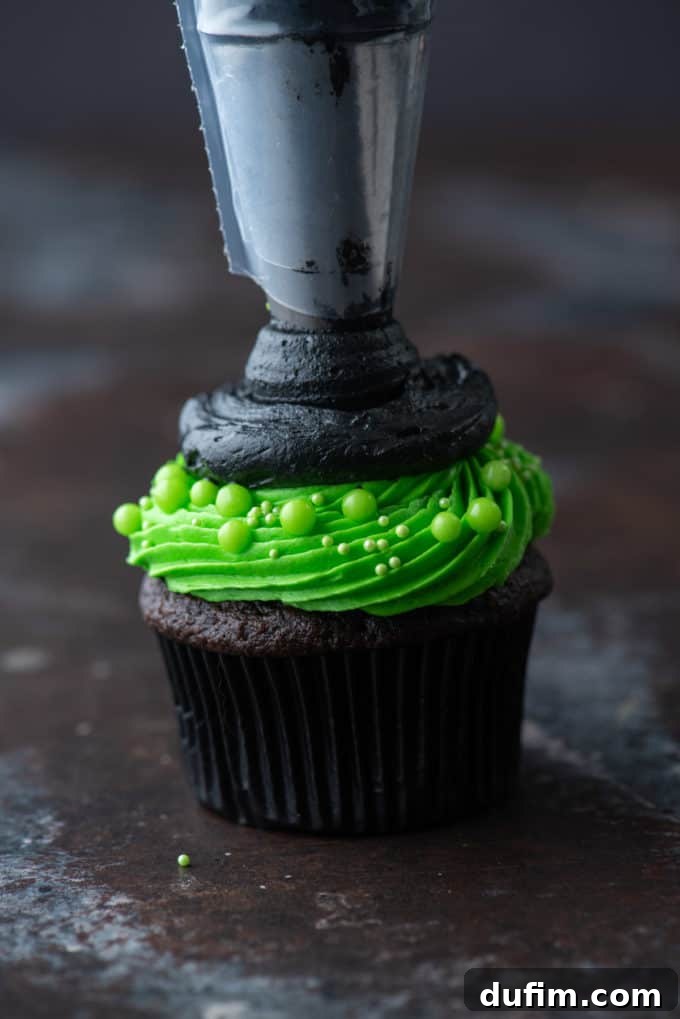 A chocolate cupcake with bright green frosting and a black frosting brim, now beginning to be piped upwards in a cone shape for the witch hat's peak.