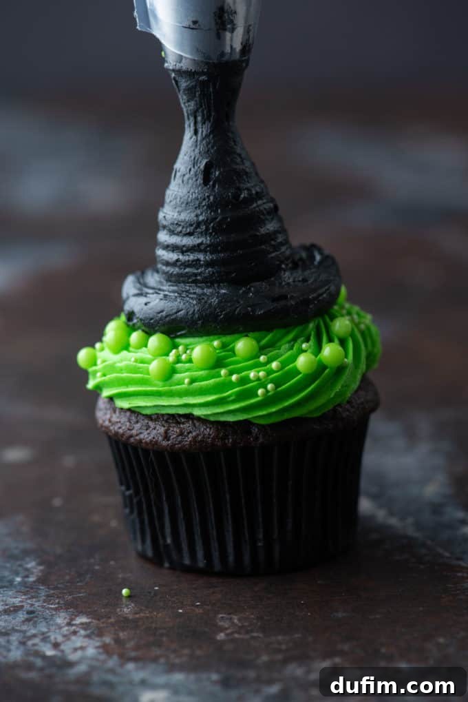 A chocolate cupcake with bright green frosting and a black frosting circle, now fully piped into a tall, pointed witch hat shape with intricate folds.