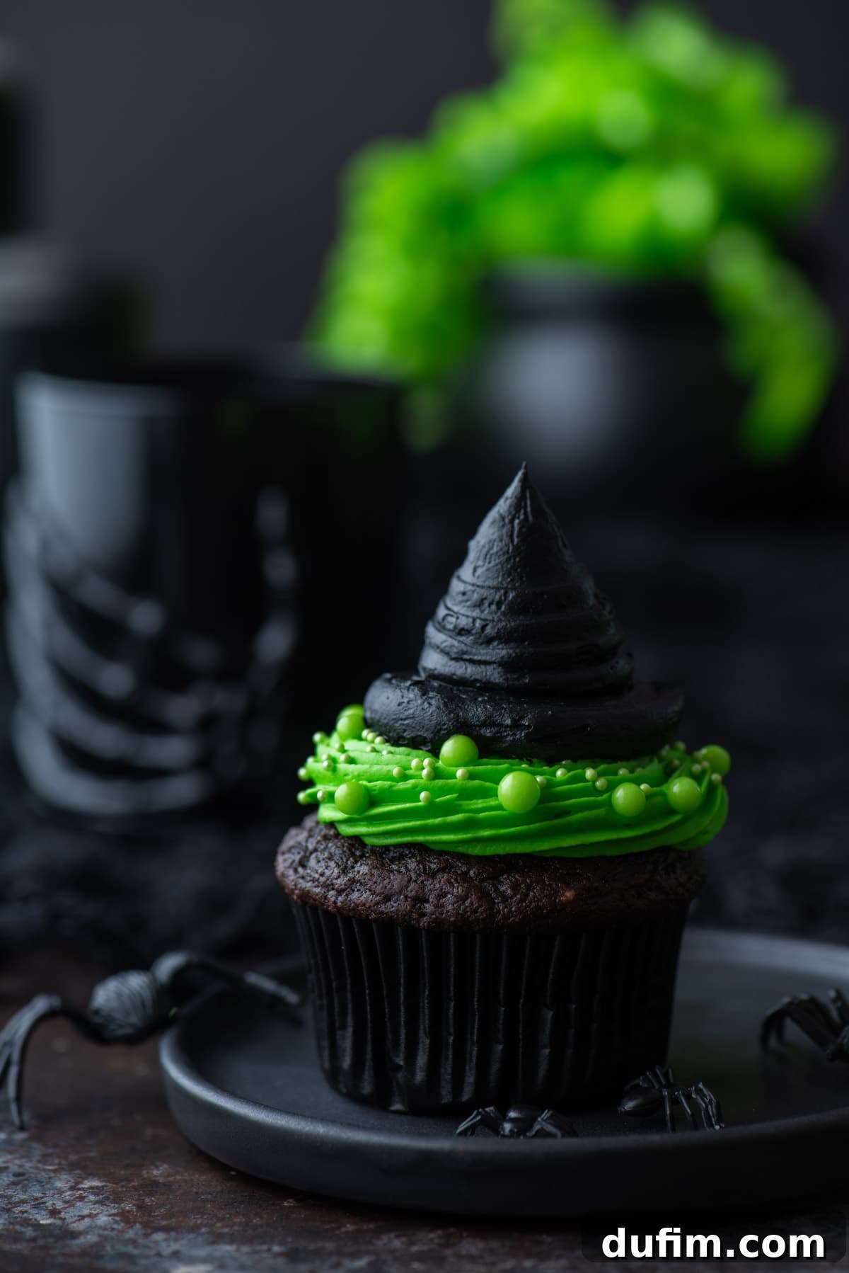 A single witch hat cupcake presented on a black plate, adorned with small black plastic spiders for an extra spooky effect.