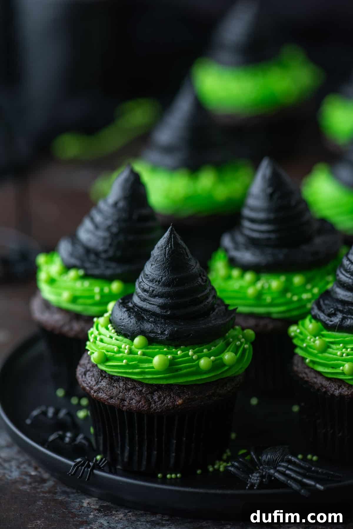 Witch hat cupcakes displayed on a black plate with small black plastic spiders scattered around for a Halloween setting.