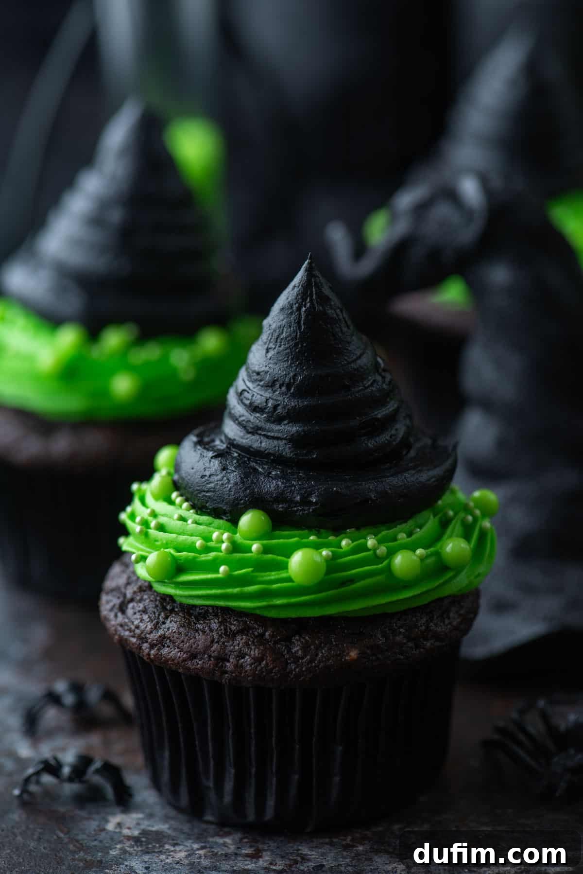 A close-up of witch hat cupcakes on a black plate, surrounded by small black plastic spiders, highlighting the detailed frosting.