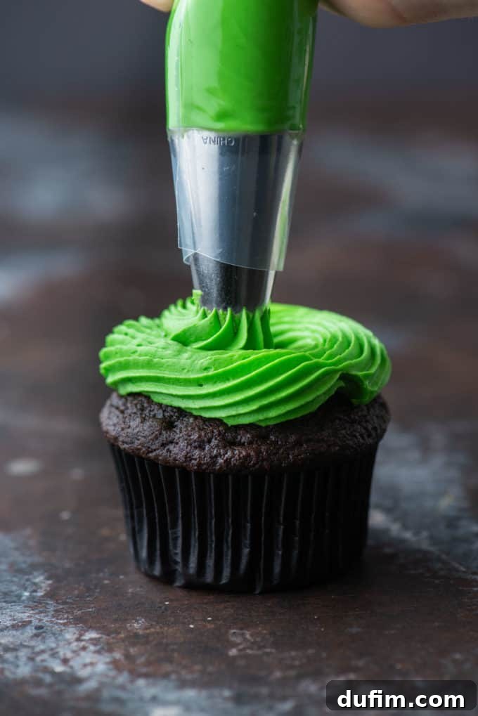 A chocolate cupcake being piped with bright green frosting in a spiral motion for the base of the witch hat.