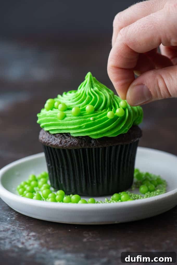 A chocolate cupcake with bright green frosting being decorated with round green sprinkles around the edges, with more sprinkles on the white plate.