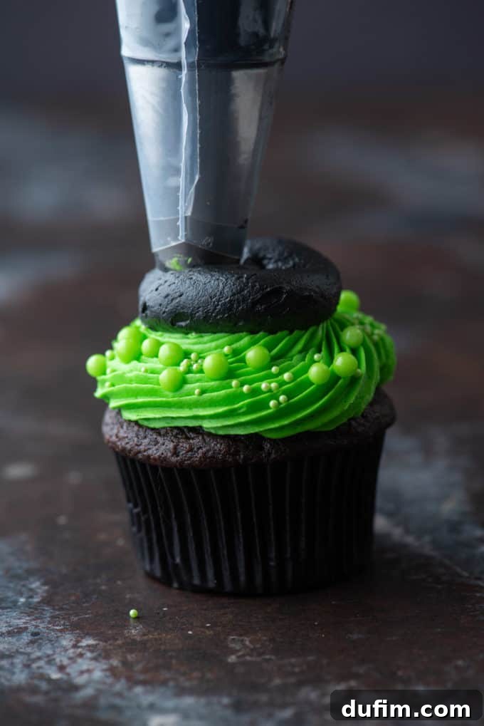 A chocolate cupcake with bright green frosting, now being piped with a wide flat circle of black frosting on top to form the brim of a witch hat.