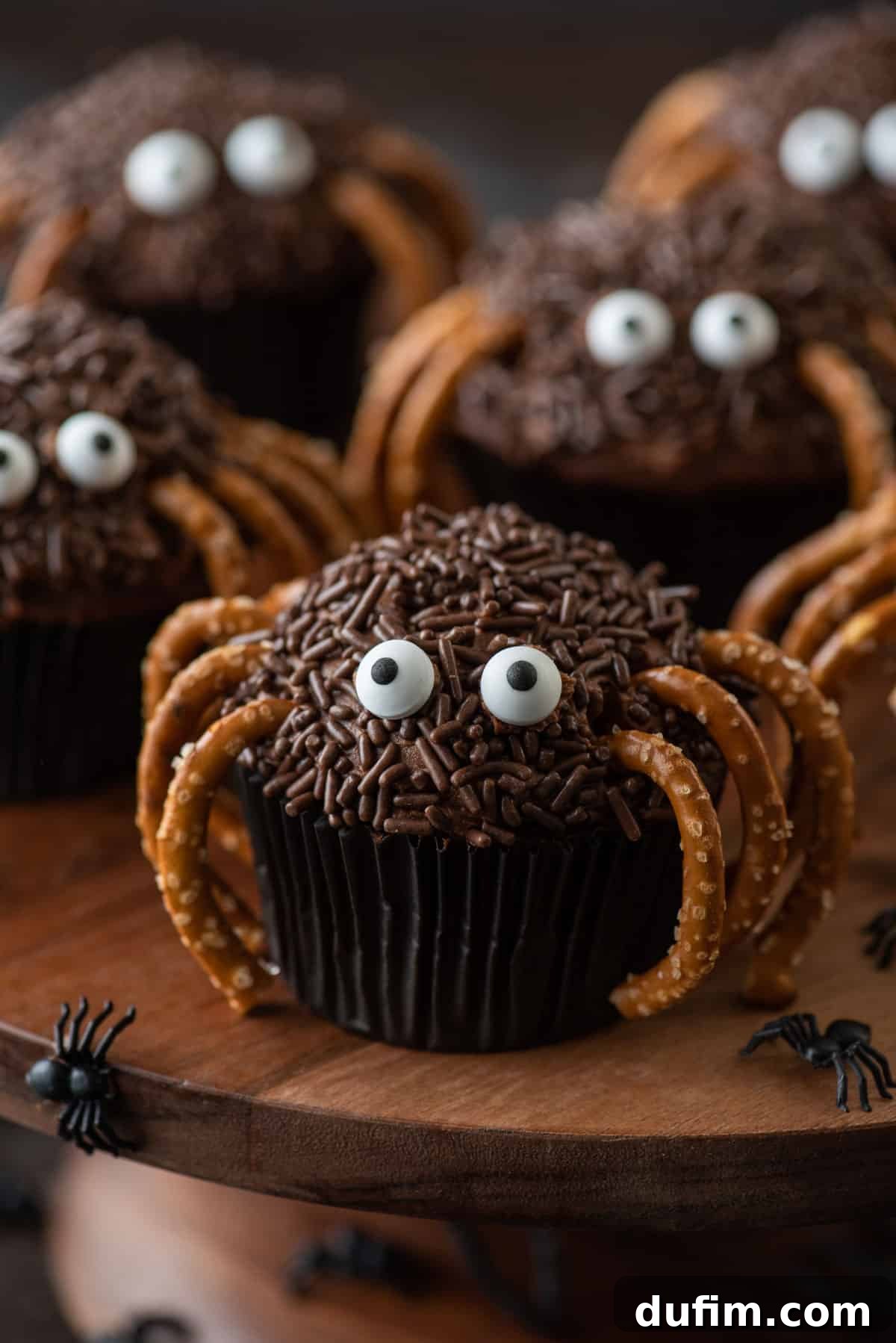 Ghoulishly Good Cupcakes 20 Playful spider cupcakes arranged on a rustic wooden surface, with scattered small black plastic spiders enhancing the Halloween theme.