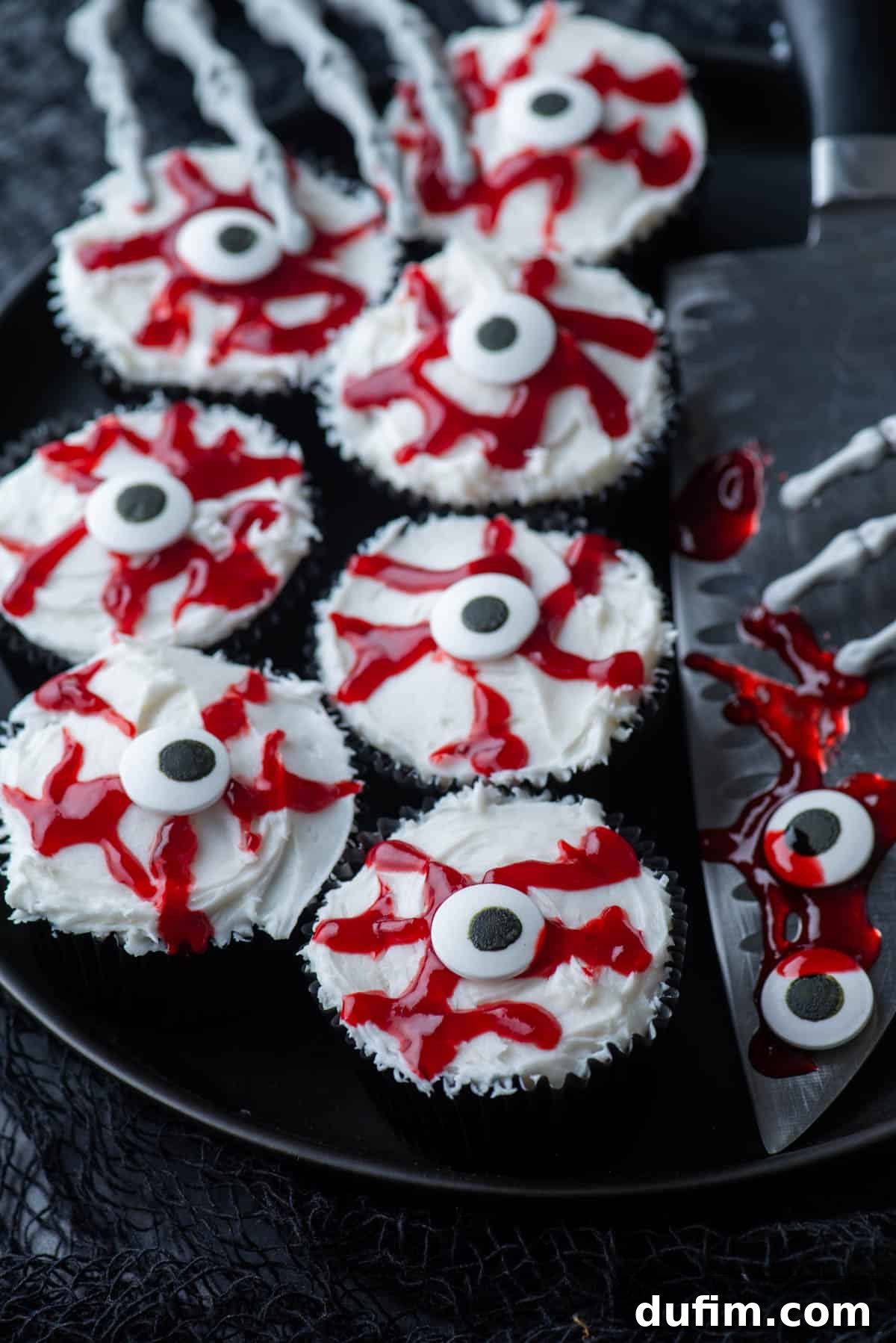 Ghoulishly Good Cupcakes 6 Horrifyingly realistic eyeball cupcakes presented on a black plate, flanked by a large knife and skeleton hands, creating a truly unsettling Halloween display.