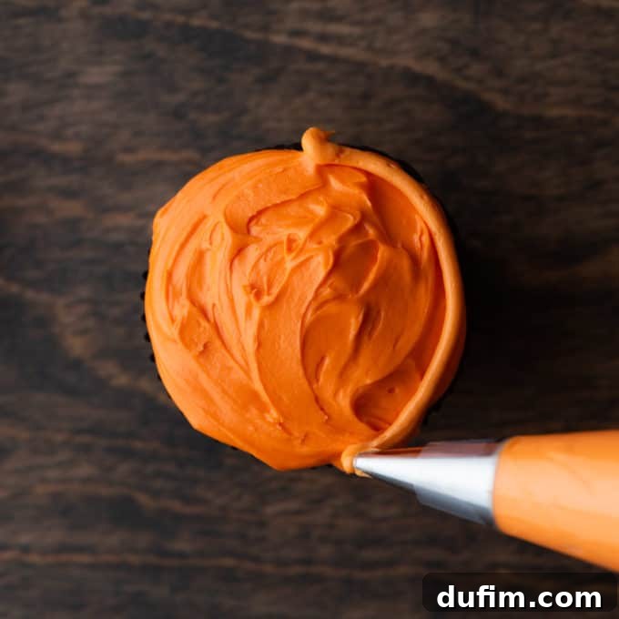 A cupcake with orange frosting, being decorated with an orange piped line along its right side to create the pumpkin's ridges.