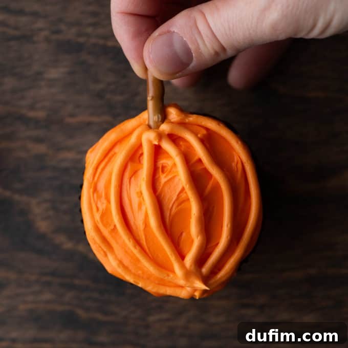 A pumpkin patch cupcake with orange frosting on a wooden surface, showing a pretzel stick being gently inserted into the top to form the pumpkin's stem.
