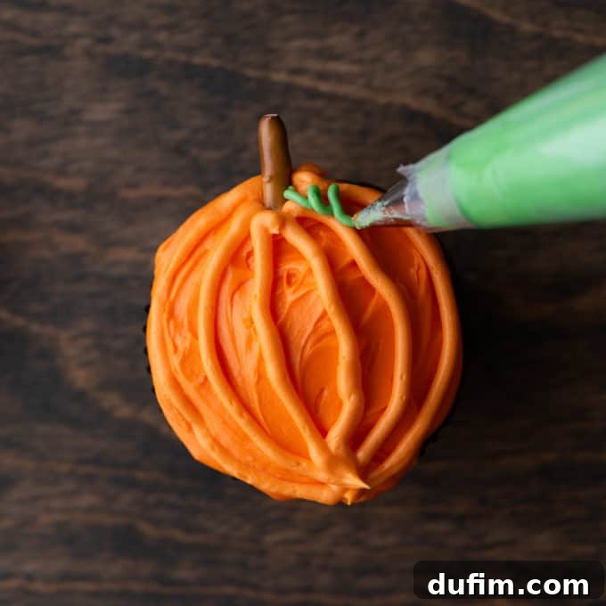 A completed pumpkin patch cupcake with orange frosting and a pretzel stick stem, being adorned with small piped green vines beside the pretzel for a realistic look.