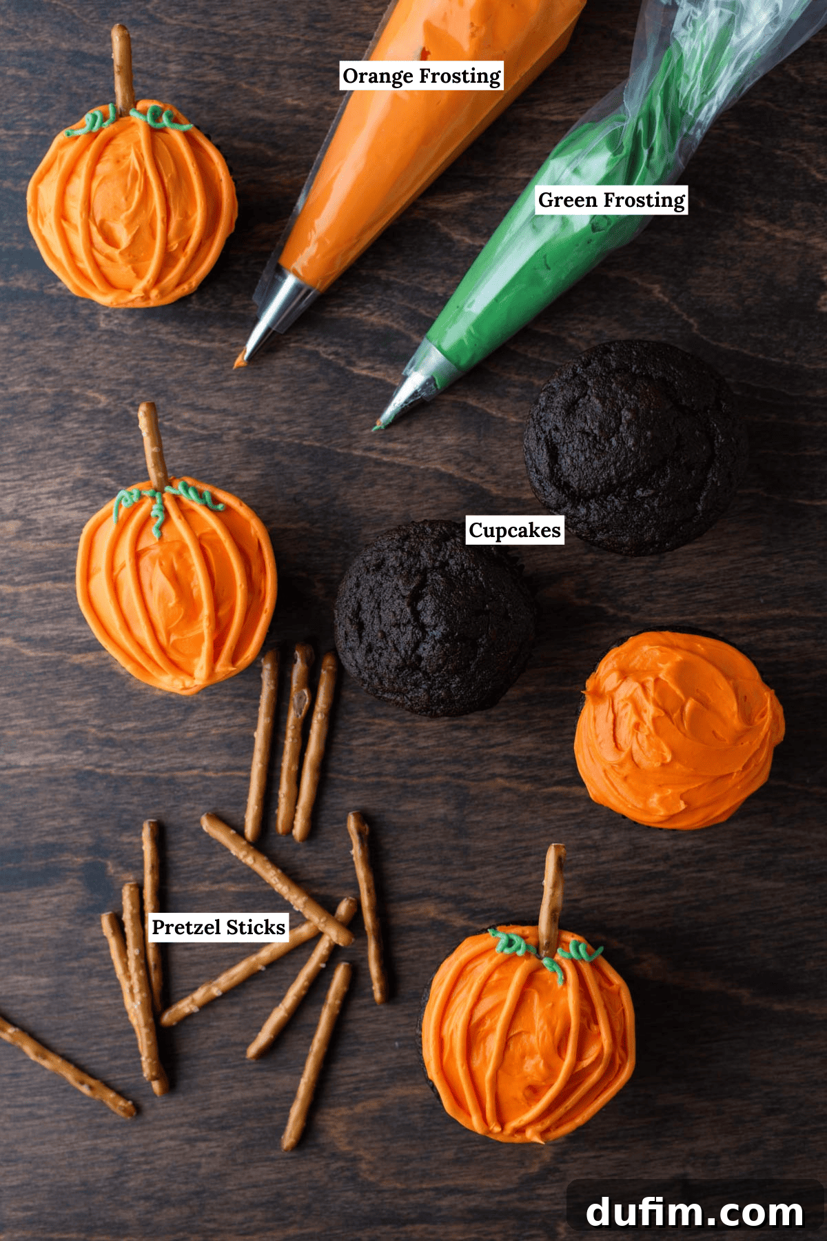 Ingredients laid out for pumpkin patch cupcakes, including vibrant green and orange frostings, baked chocolate cupcakes, and pretzel sticks, with a few finished cupcakes scattered around.