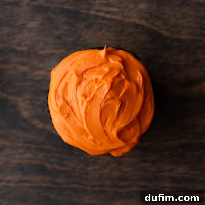 A freshly frosted cupcake with smooth orange frosting, ready for the next decorating steps, sitting on a wooden surface.