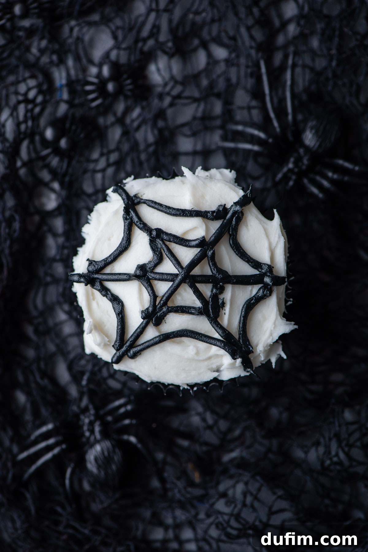 An almost completed black spider web design piped onto a white-frosted cupcake, showcasing the delicate and spooky pattern.