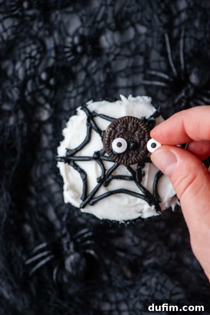 Attaching small candy eyeballs to the top of a mini Oreo with a dab of frosting, transforming it into a cute spider for the cupcake decoration.