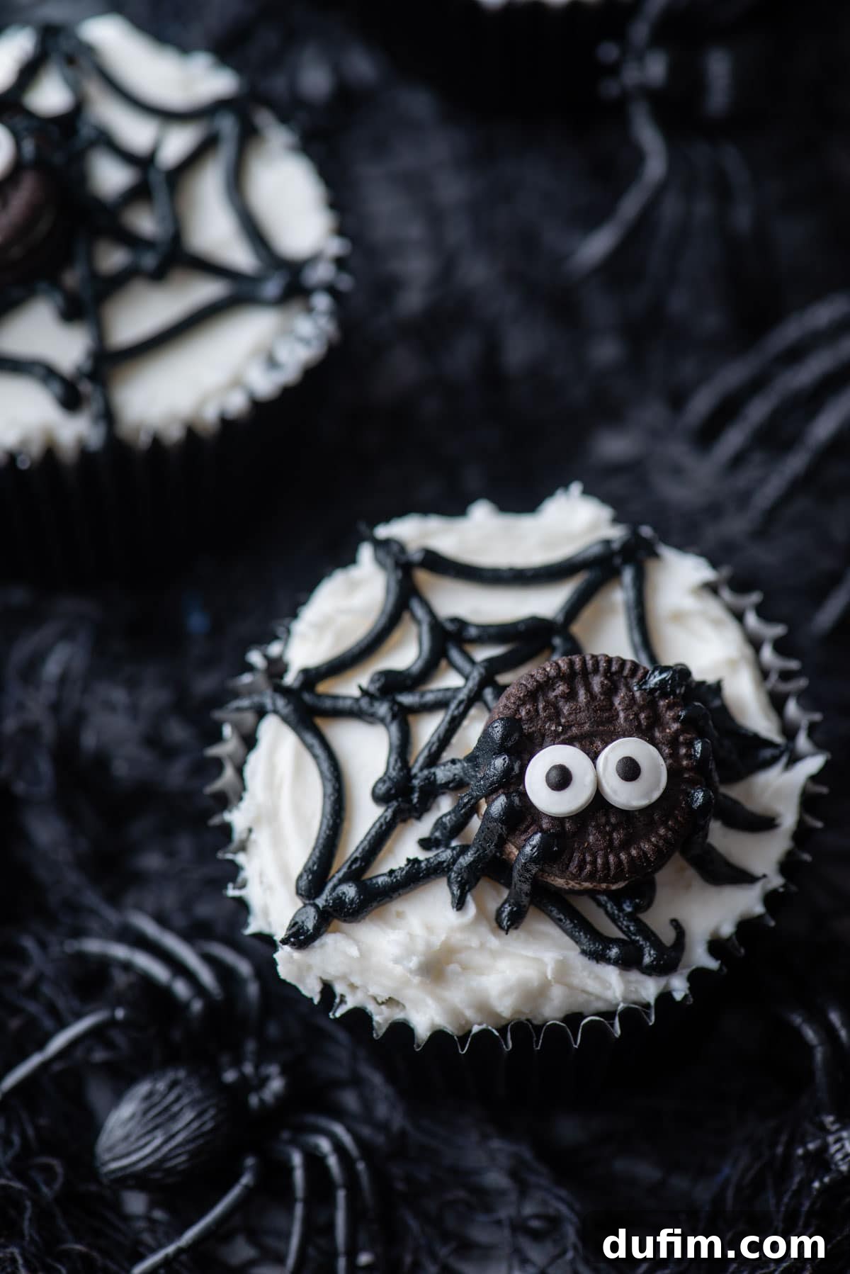 A single spider web cupcake displayed on black netting, with additional cupcakes and small black plastic spiders creating a spooky background.
