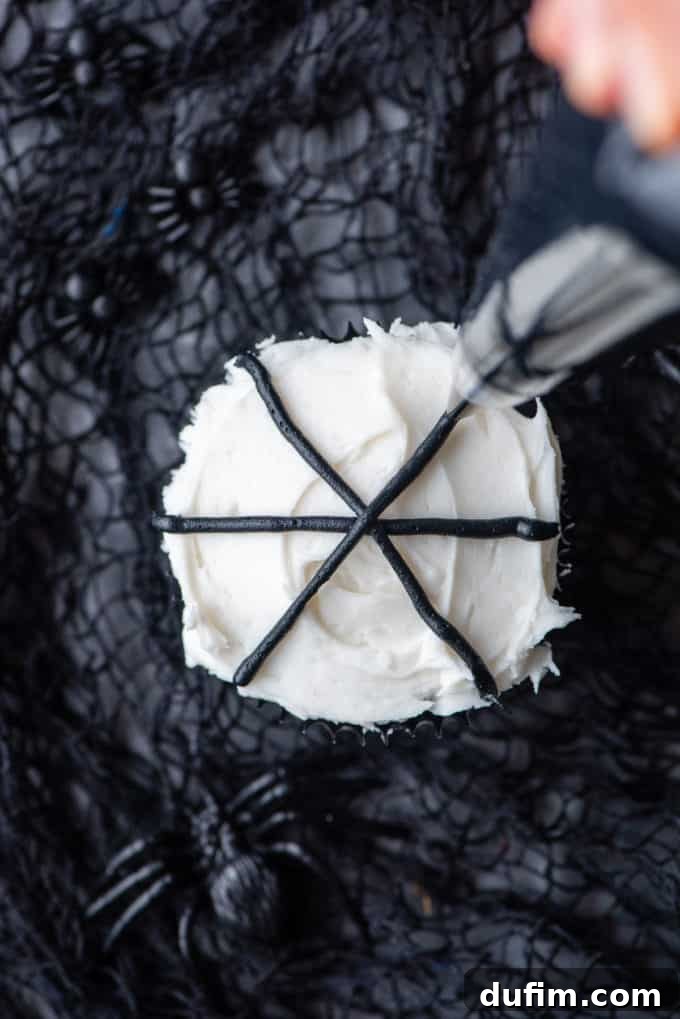 Piping a straight black line across a white-frosted cupcake, the initial step in creating a spider web decoration.