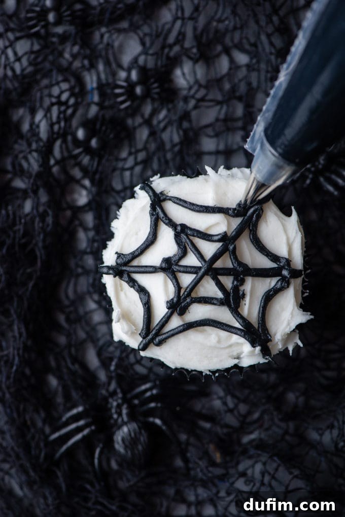 The process of piping intricate black spider web lines onto a cupcake that has been smoothly frosted with white icing.