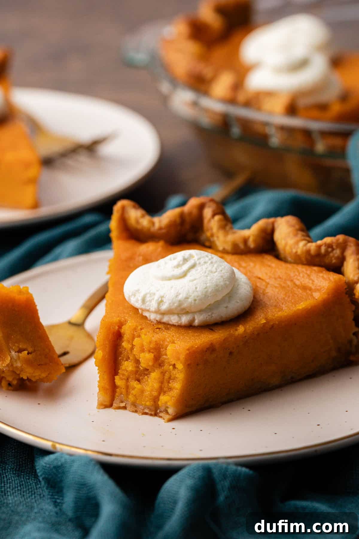 Grandma's Secret Sweet Potato Pie Recipe 3 a slice of sweet potato pie on a white plate with one bite taken out on a fork laying beside the pie on the plate. The plate is sitting on a dark teal towel and there is another plate with a pie slice and the glass pie dish of pie in the background