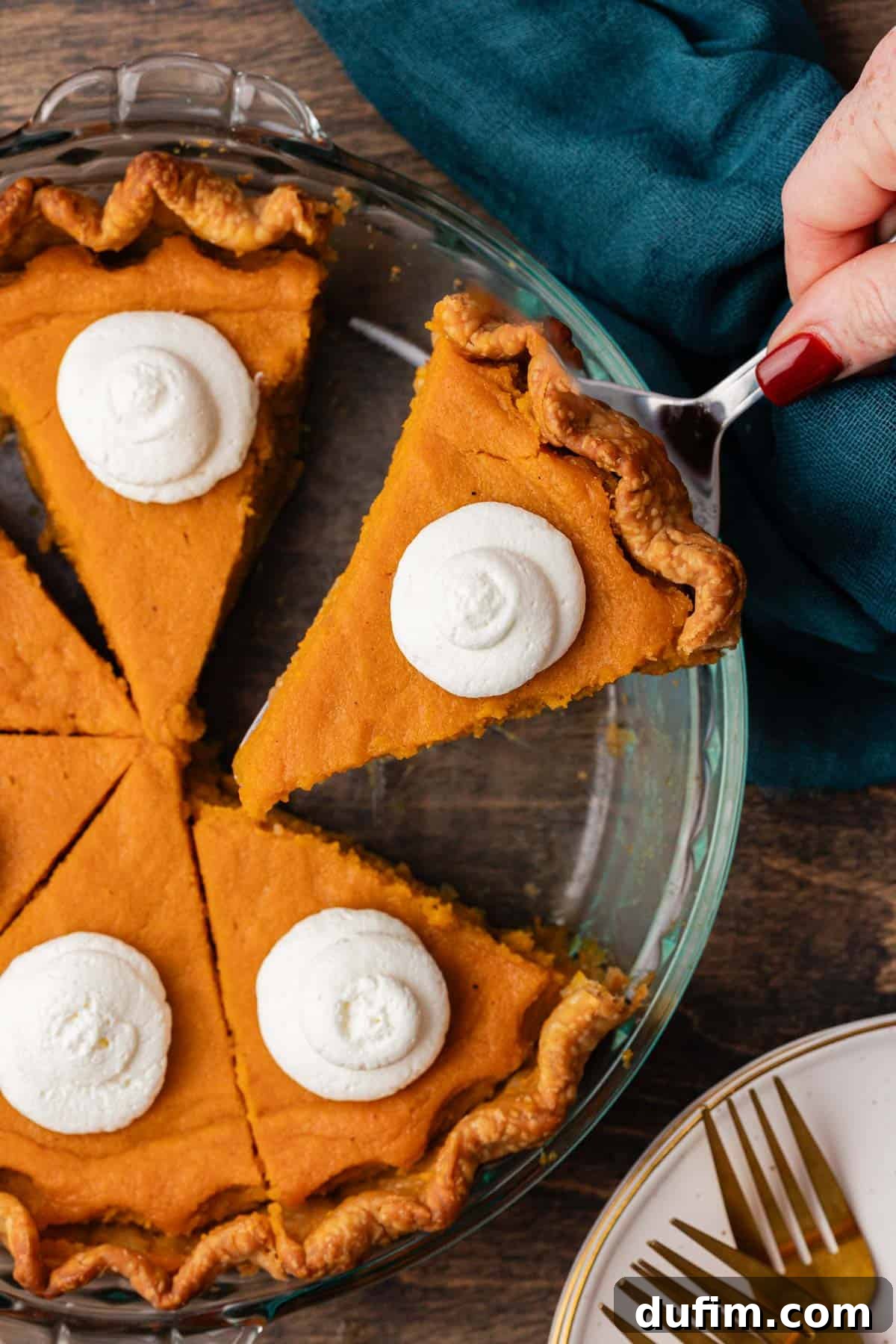 Grandma's Secret Sweet Potato Pie Recipe 6 a glass pie dish with sweet potato pie slices topped with dollops of whipped cream, with one slice being lifted out of the pie dish and a dark teal towel beside it