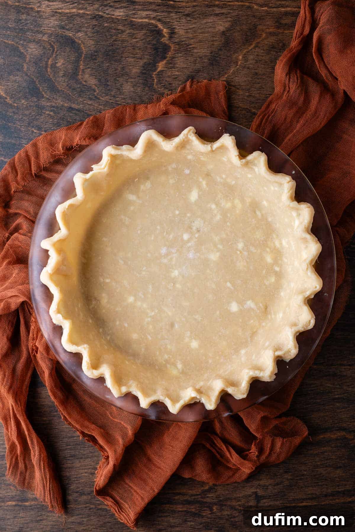A perfectly baked gluten-free pie crust in a glass pie dish, resting on a dark orange cloth on a rustic wood surface, ready to be filled.