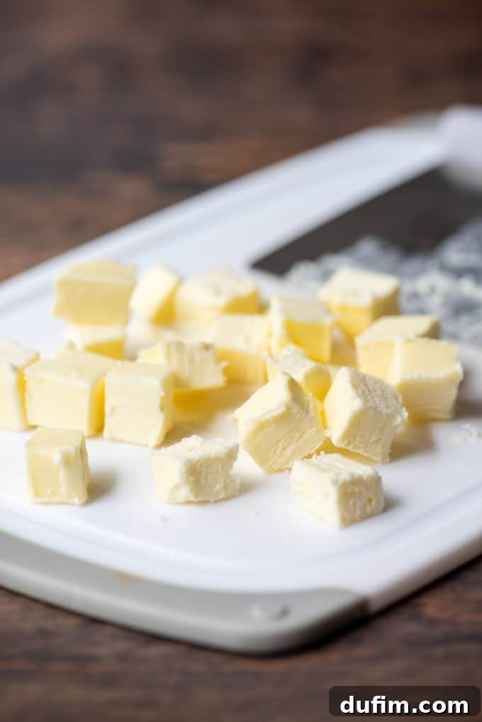 Small, uniformly cut cubes of cold butter and a pastry cutter resting on a white cutting board, set against a warm wood surface, illustrating chilled ingredients.