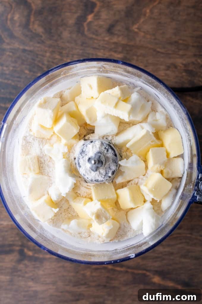 Dry pie crust ingredients in a food processor bowl on a wooden surface, with cubes of butter and shortening waiting to be mixed in.