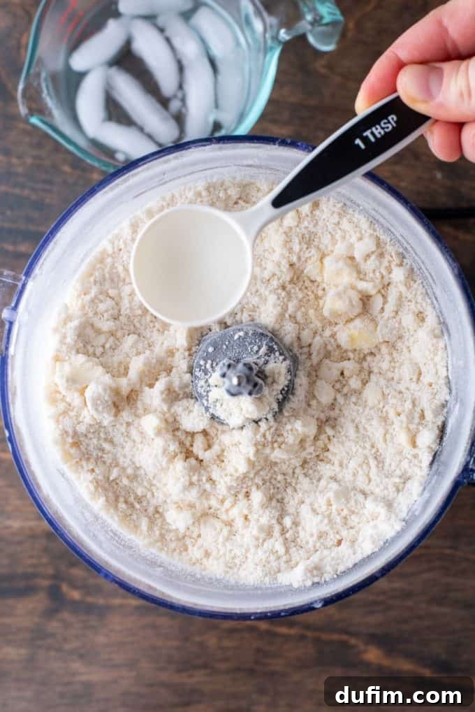 Gluten-free pie crust ingredients combined in a food processor, with a measuring cup of ice water nearby, and a tablespoon of water about to be added.