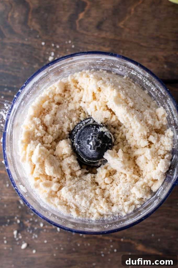 Gluten-free pie crust dough, still somewhat crumbly but coming together, mixed in a food processor bowl on a wooden surface with scattered flour and dough specs.