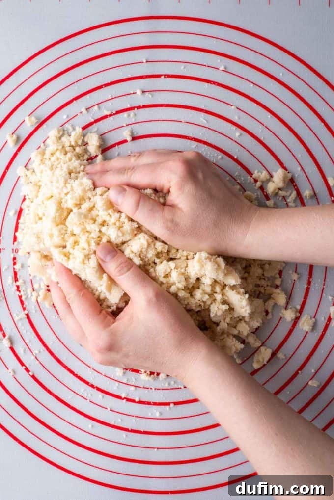 Crumbly chunks of gluten-free pie crust dough on a red and white pie mat, with two hands gently gathering and starting to form the dough into a disc.