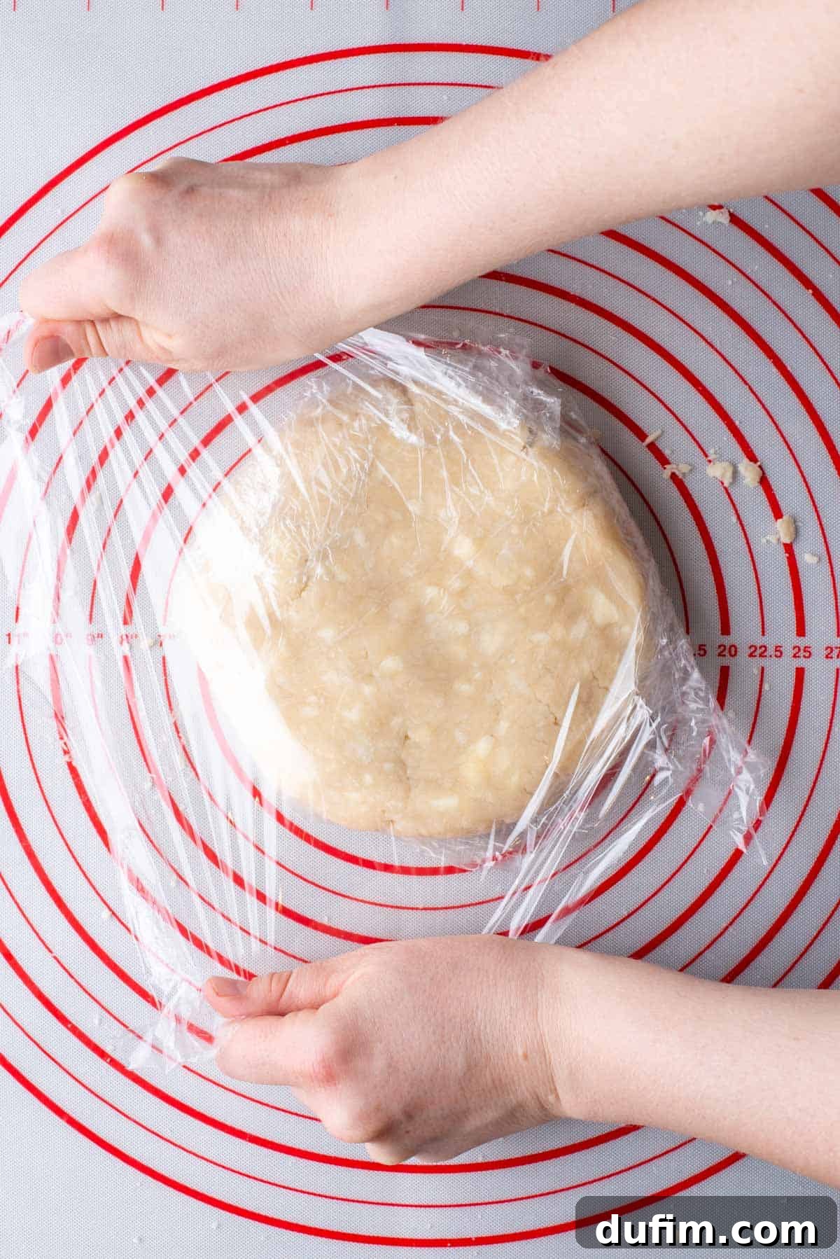 A disc of perfectly shaped gluten-free pie crust dough being meticulously wrapped with clear plastic wrap on a red and white pie mat, ready for chilling.