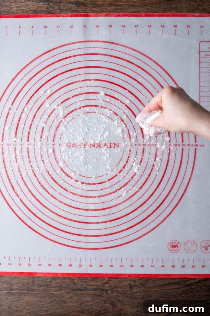 A hand expertly dusting a red and white pie mat, resting on a wooden surface, with gluten-free flour to prepare for rolling dough.