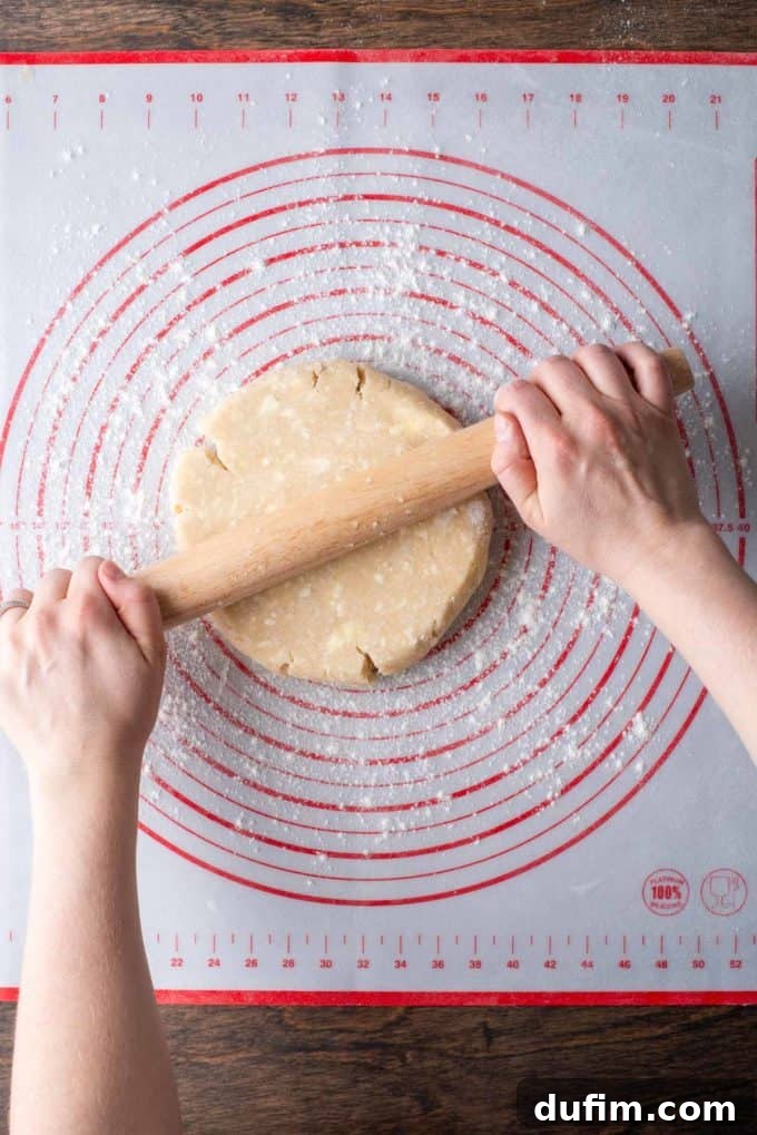A disc of gluten-free pie crust dough being gently rolled with a wooden rolling pin on a floured red and white pie mat, demonstrating initial rolling technique.