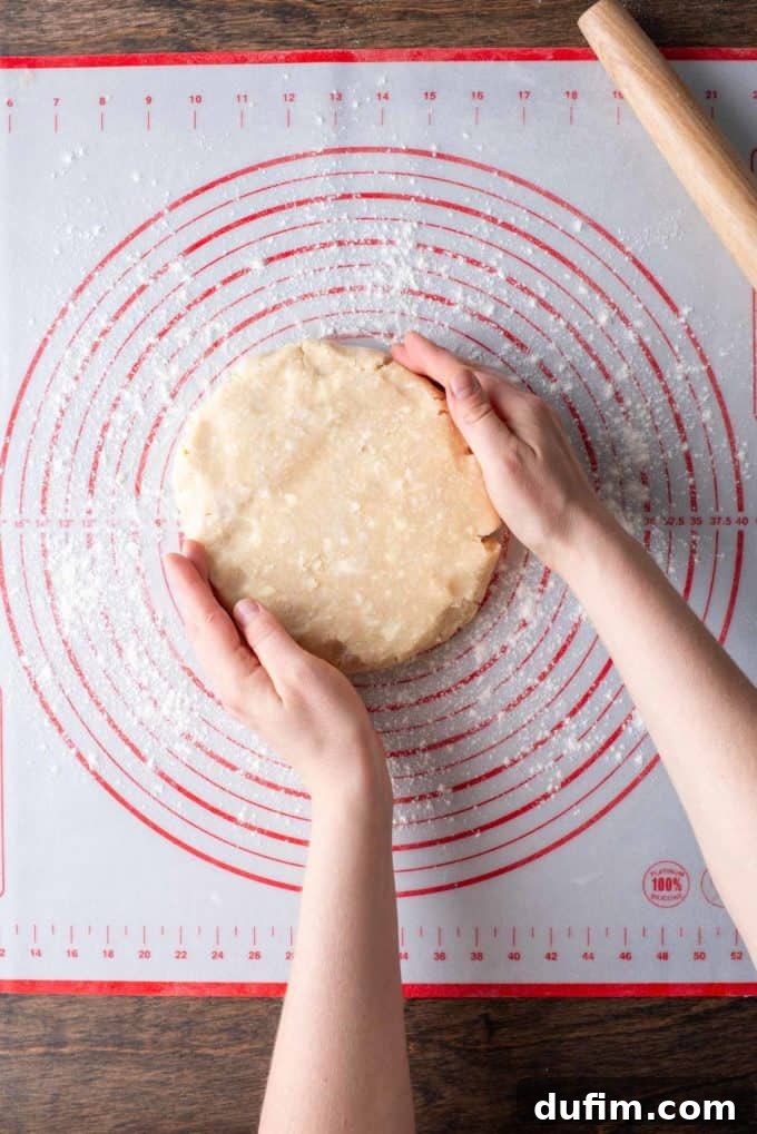 A disc of gluten-free pie crust dough being carefully placed on a floured red and white pie mat, with a wooden rolling pin at the side, setting up for rolling.