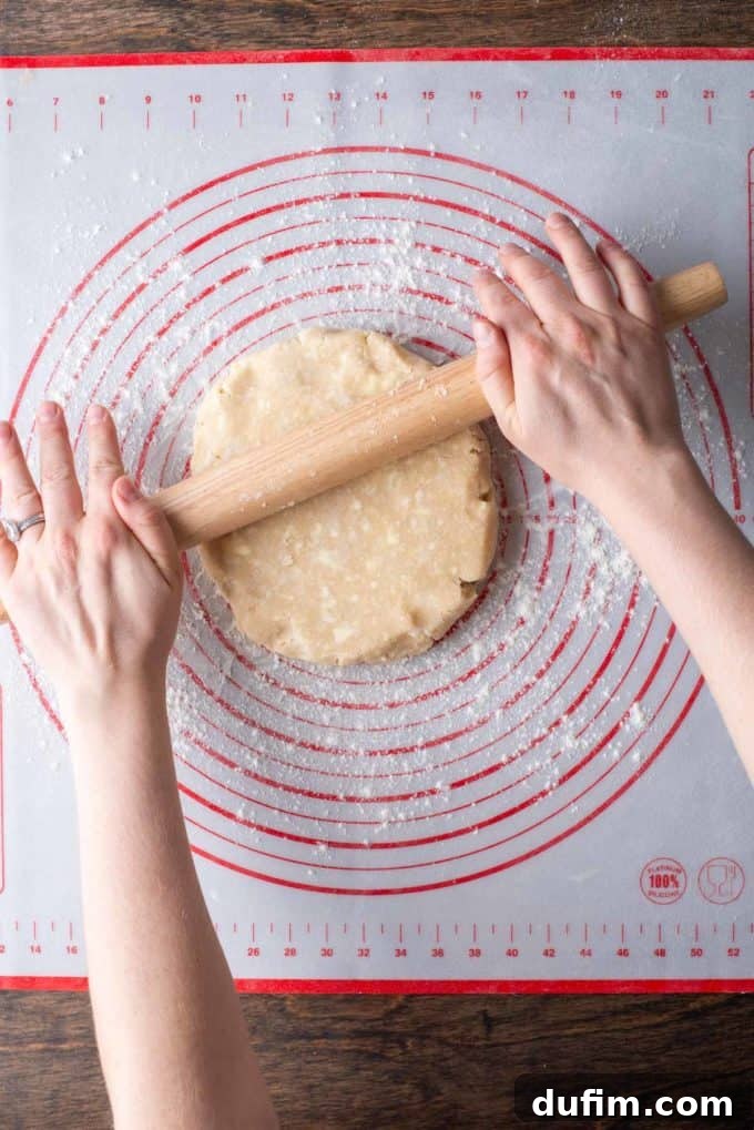 A disc of gluten-free pie crust dough being placed on a floured red and white pie mat on a wooden surface, with a wooden rolling pin positioned nearby, ready to begin rolling.
