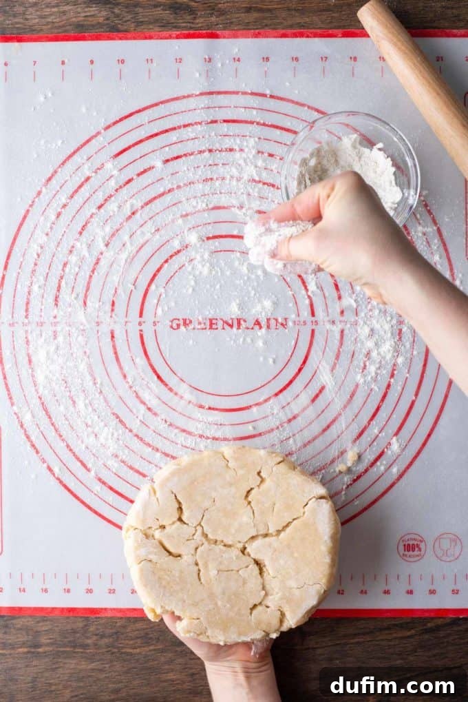 A disc of pie dough on a red and white pie mat being lightly sprinkled with flour from a small glass bowl, with a wooden rolling pin to the side, maintaining non-stickiness.
