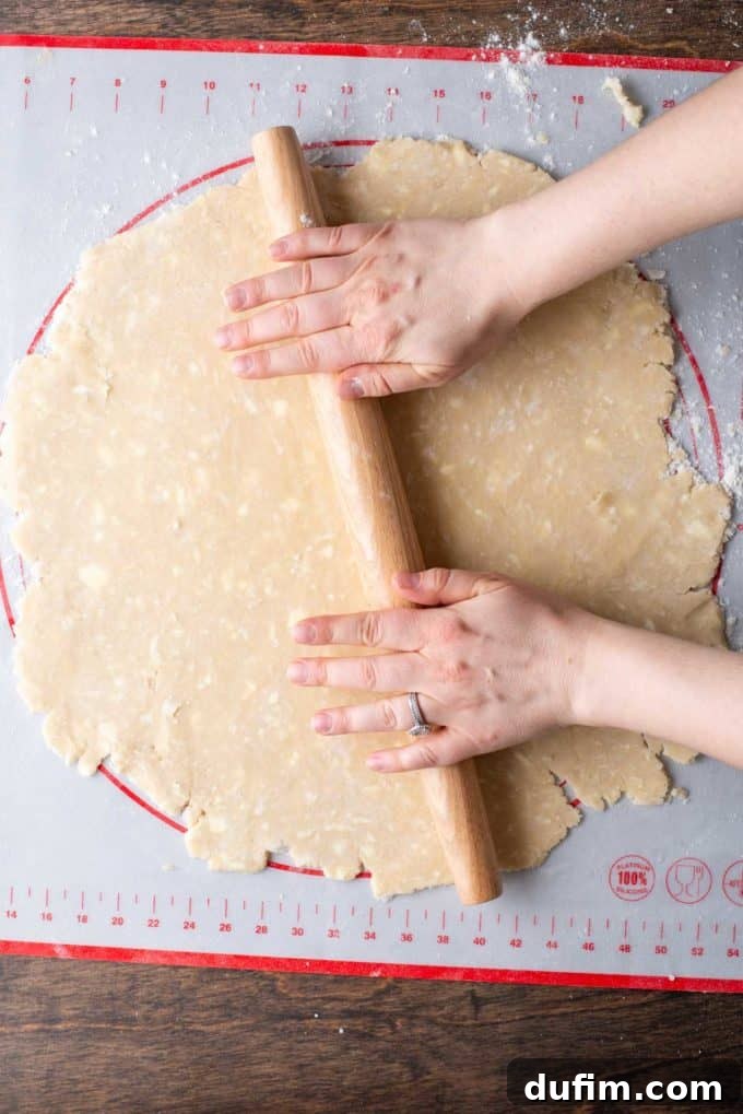 A large, somewhat imperfect but expanding circle of gluten-free pie crust dough being rolled out with a wooden rolling pin on a floured red and white pie mat.