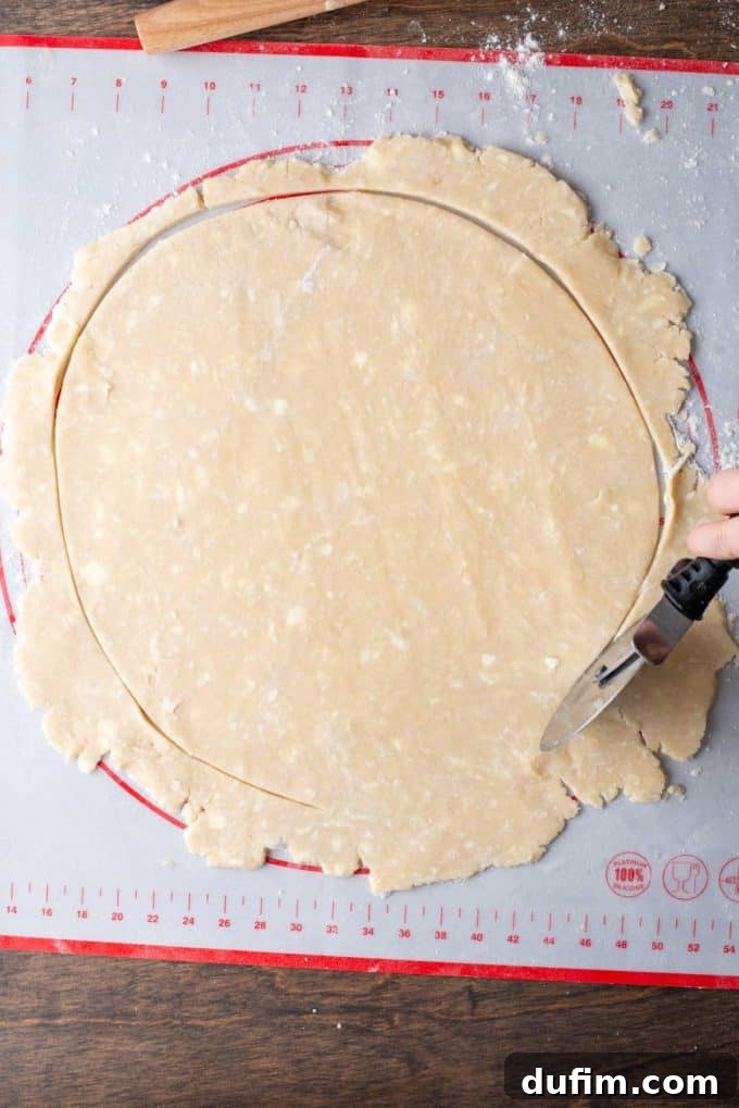 A large, circular piece of gluten-free pie crust dough being trimmed with a pizza cutter on a floured red and white pie mat to achieve a perfect round shape.