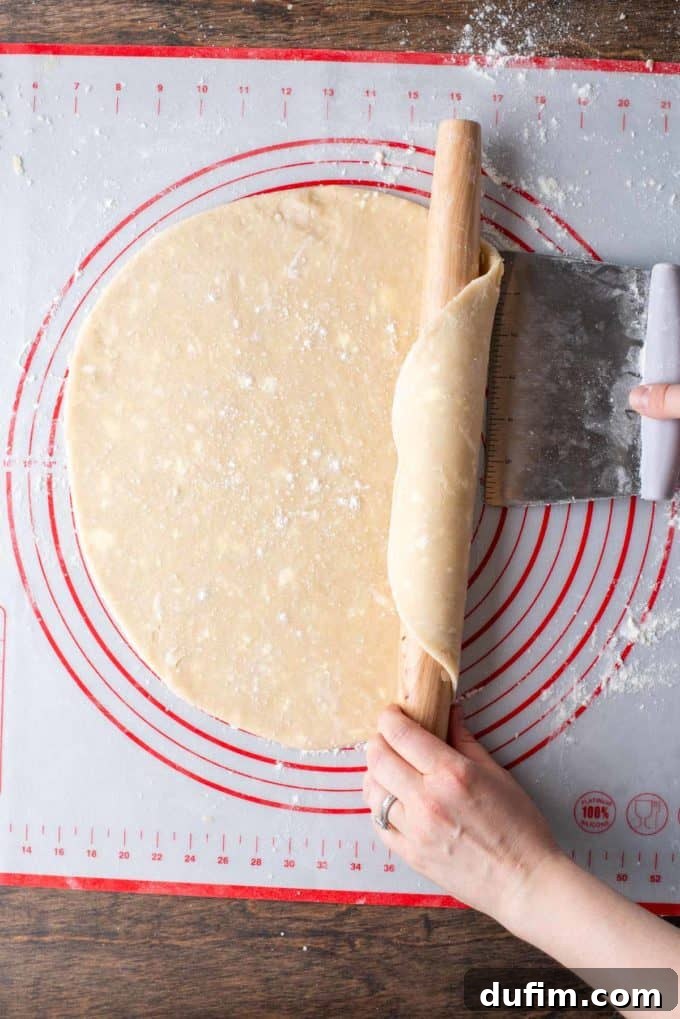 Rolled out gluten-free pie crust dough being carefully lifted by a pastry cutter and rolled onto a wooden rolling pin, preparing for transfer.
