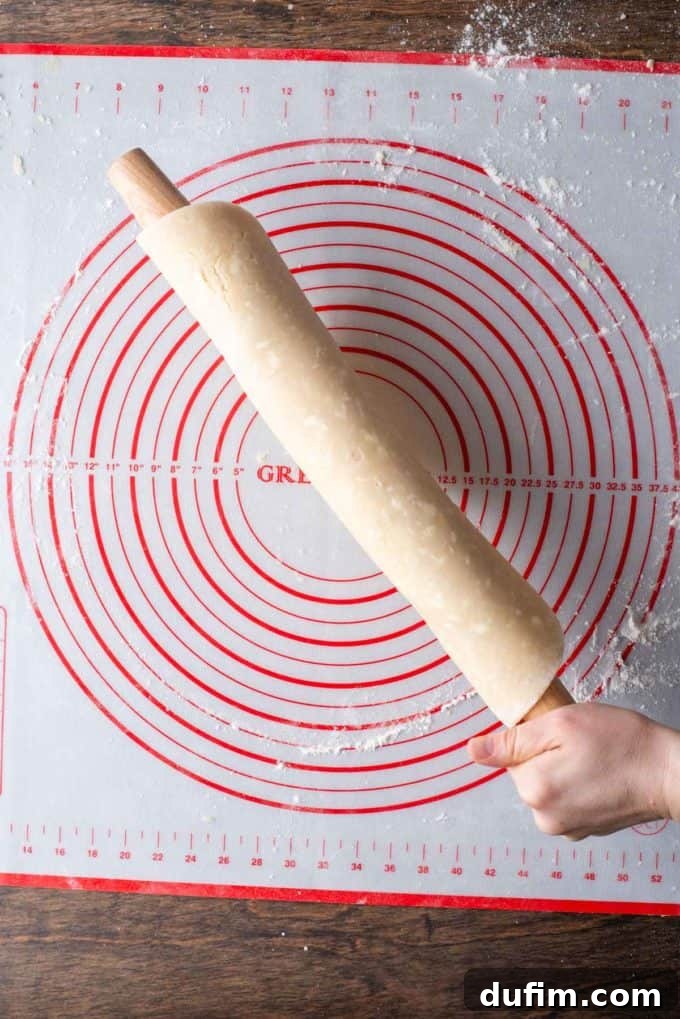 Trimmed, rolled-out gluten-free pie crust dough, fully wrapped around a wooden rolling pin, held over a floured red and white pie mat, ready for placement.
