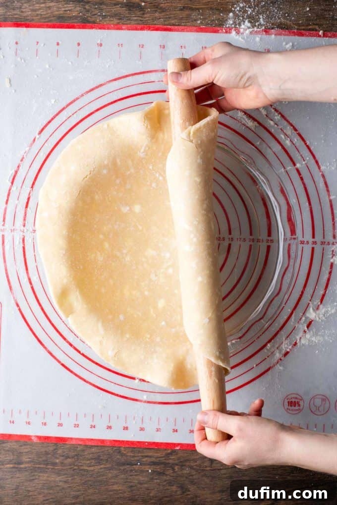 Gluten-free pie crust being gently unrolled from a wooden rolling pin directly into a glass pie dish, positioned on a red and white pie mat, ensuring even coverage.