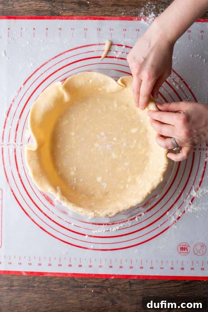 The edges of the gluten-free pie crust being neatly folded over the rim of a pie dish, which sits on a red and white pie mat, preparing for crimping.