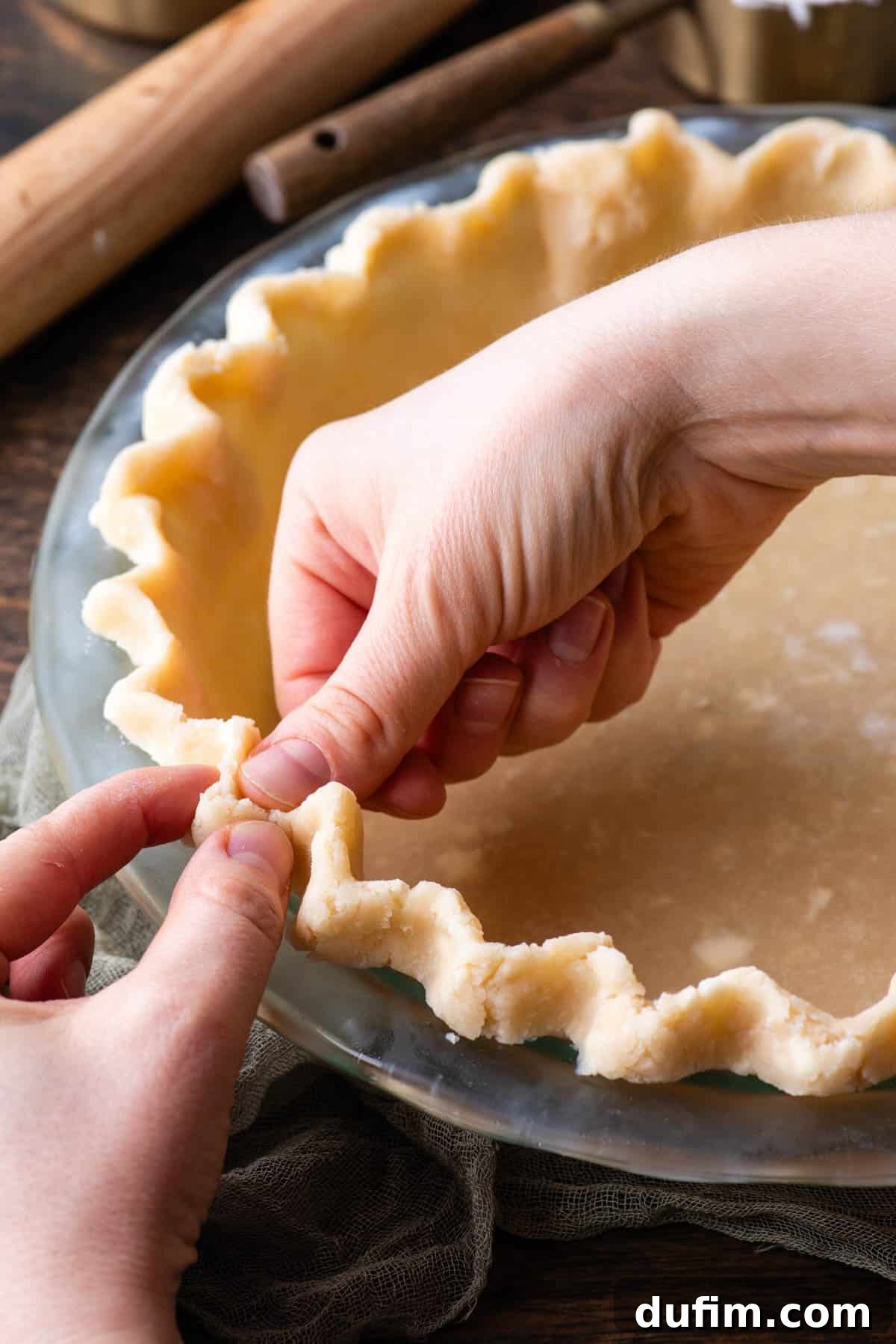 Two hands delicately crimping the edge of a gluten-free pie crust in a clear glass pie pan, demonstrating the fluting technique.