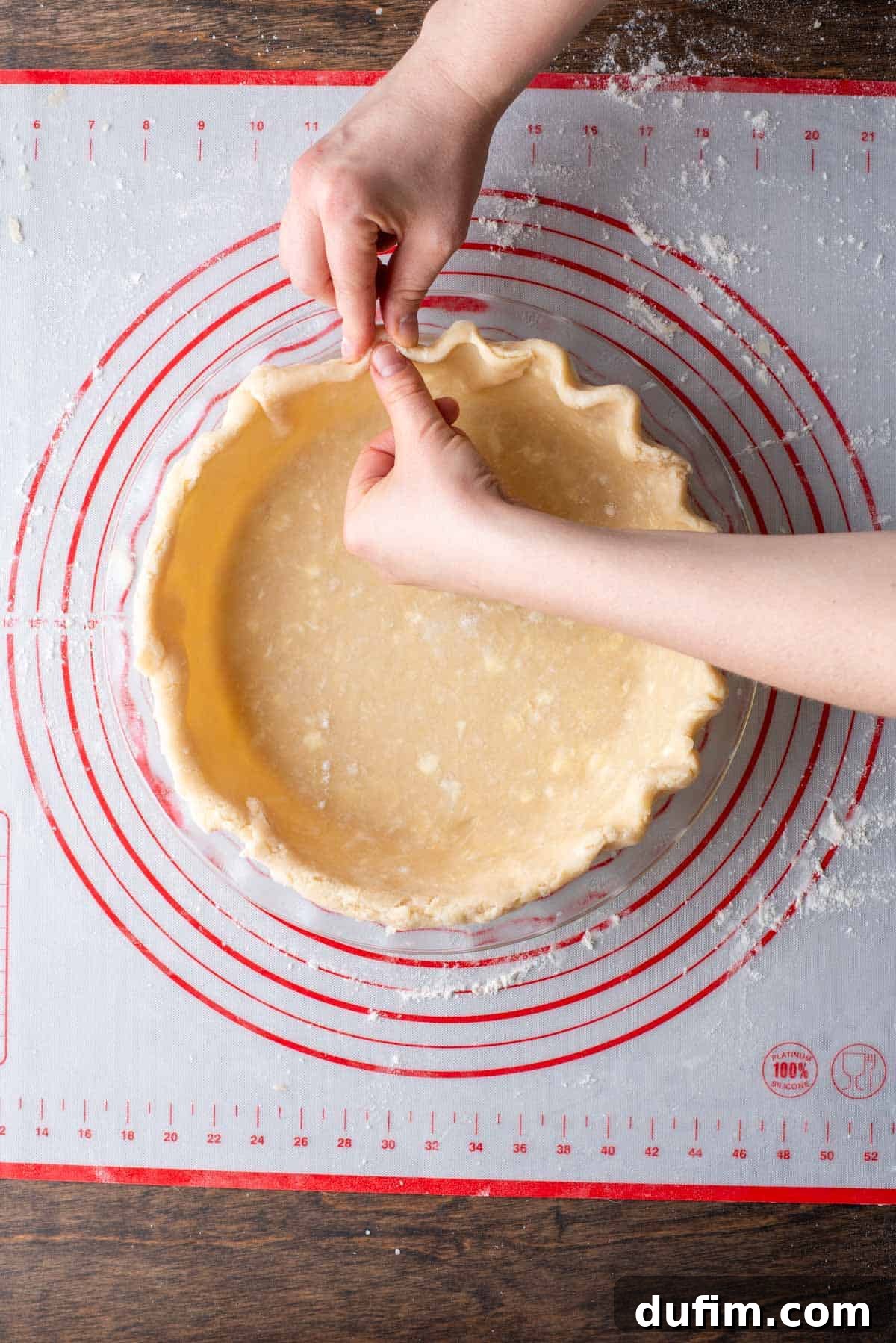 A gluten-free pie crust being meticulously crimped around the edges of a glass pie dish, resting on a floured red and white pie mat, highlighting the fluting technique.